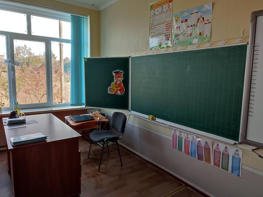 Класна кімната зі сторони вчительського місця. Два столи та стілець. На одному зі столів лежить складений ноутбук. На стіні праворуч - класна дошка зеленого кольору. Над дошкою – навчальні таблиці. Під дошкою — декор у вигляді олівців.