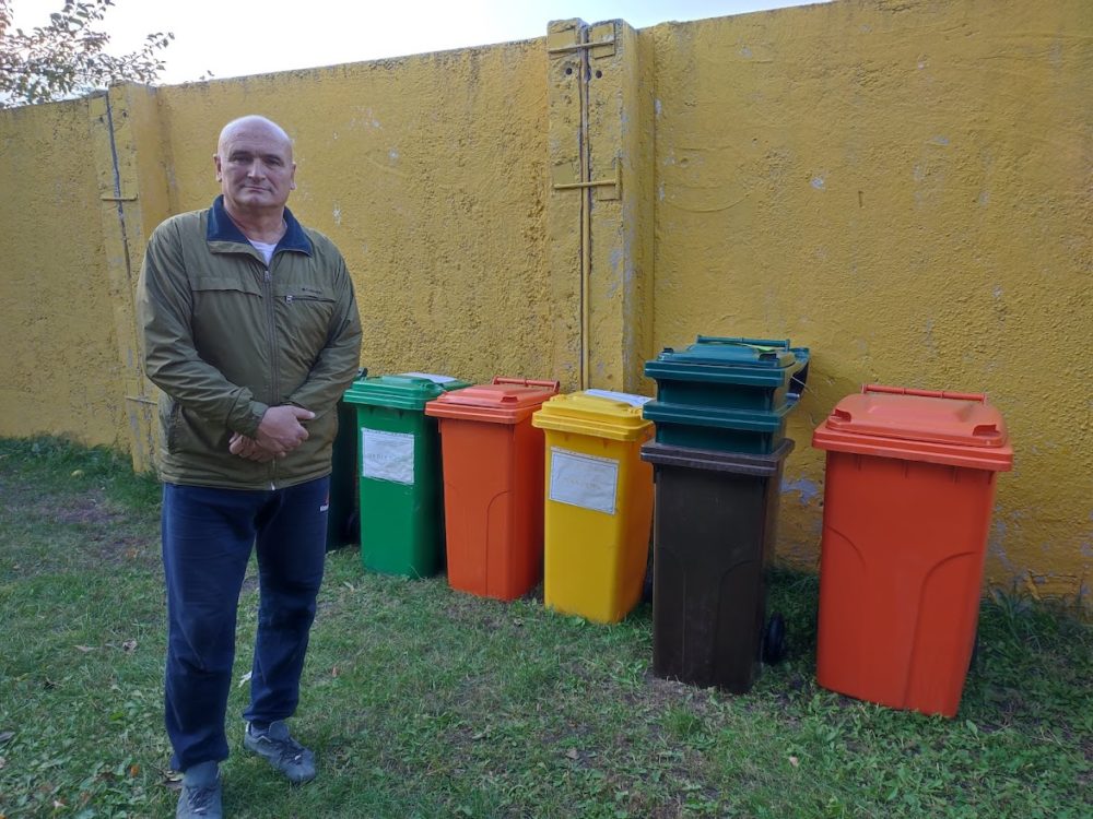 Чоловік середнього віку стоїть на тлі різноколірних сміттєвих контейнерів.