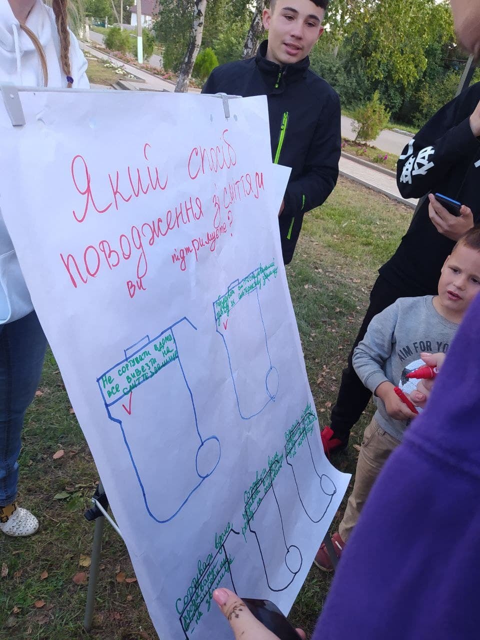 Двоє підлітків тримають плакат з написом «Який спосіб поводження зі сміттям ви підтримуєте?», пропонуючи жінці обрати варіант відповіді.