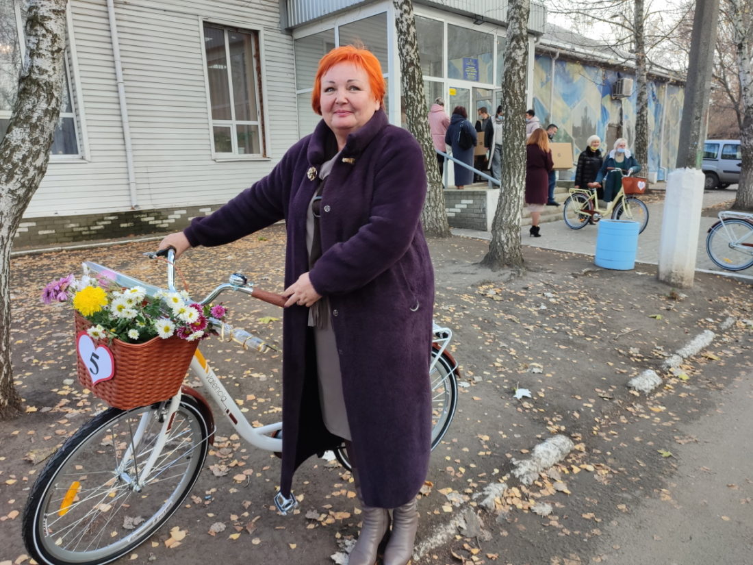 Ошатно вдягнена жінка тримає велосипед. Спереду велосипеда кошик з квітами. На кошику - паперове серце з цифрою 5