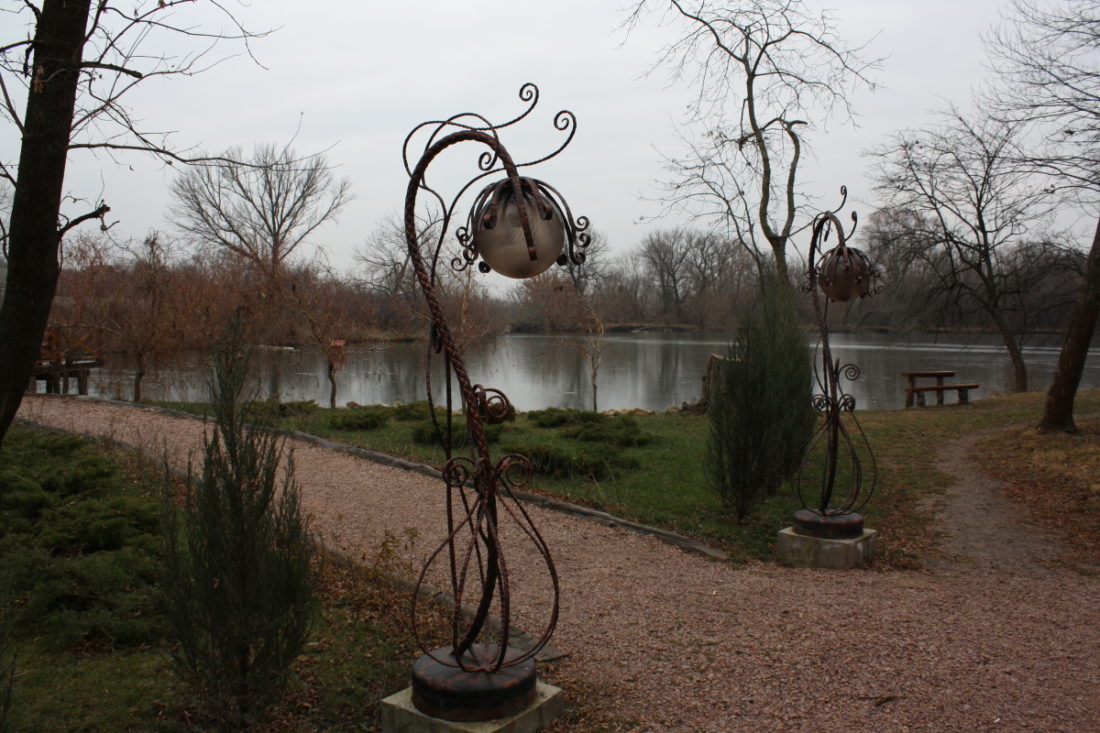 Гравієва доріжка з двома ліхтарями у псевдомодернистському стилі веде до озера, на березі якого розташовані столики і лавки.