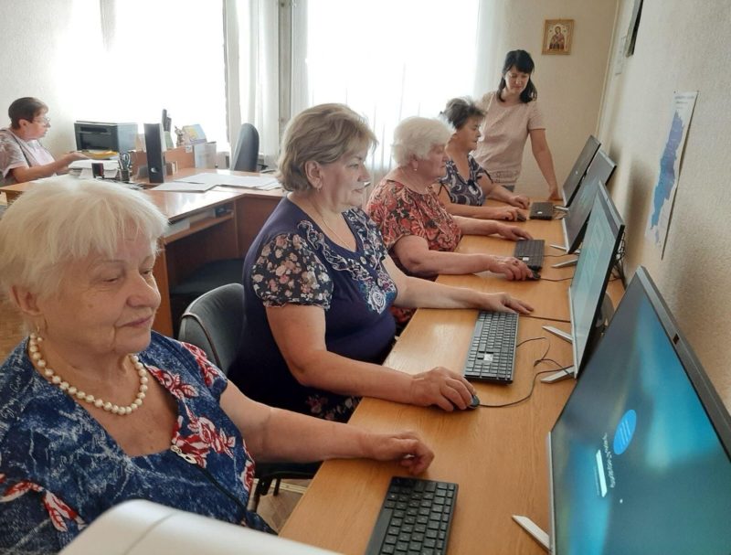 Чотири жінки похилого віку сидять за комп’ютерами. На задньому плані стоїть молодша жінка, яка, можливо контролює процес