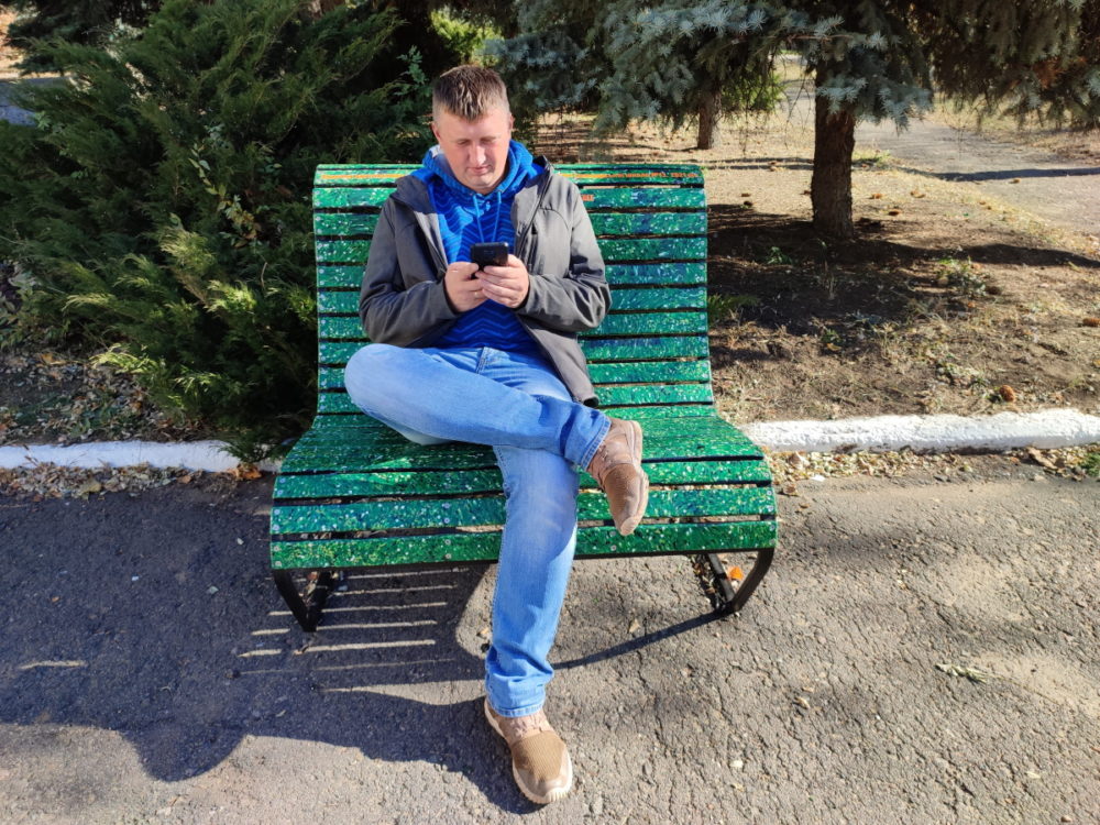 Михайло Кулішов сидить на зеленій пластиковій лаві й дивиться на екран смартфона.