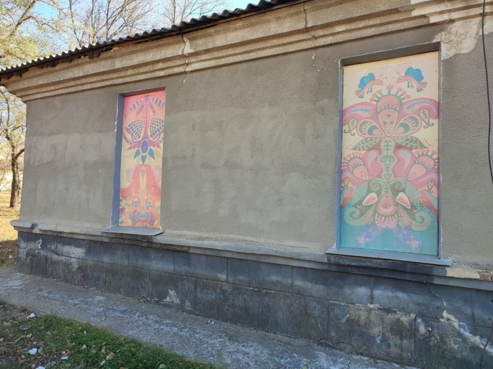 Стіна будівлі з двома вікнами. У заколочених віконницях рослинні та тваринні зображення виконані у стилі примітивізму.