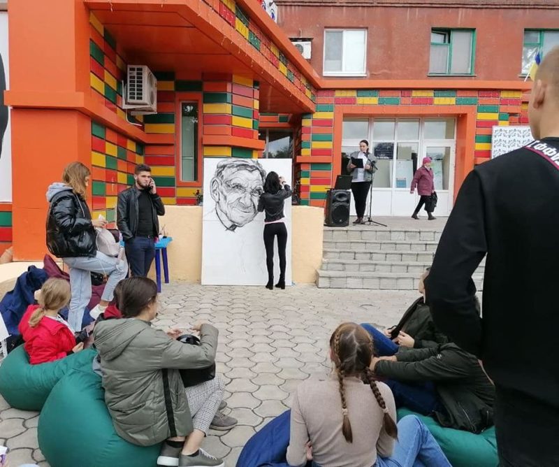 Група людей спостерігає за дівчиною, яка малює портрет на площині розміром приблизно метр на два. На задньому плані сходи до будівлі. На ґанку – акустична система і жінка біля мікрофона.