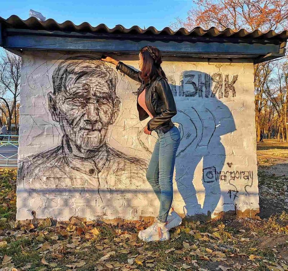 Дівчина на стіні автобусної зупинки малює чорним кольором портрет немолодого чоловіка. Поруч напис – «Залізняк».
