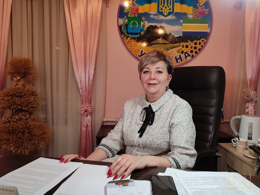 Голова Олександрівської громади Людмила Борисевич сидить за столом у кабінеті на тлі стенду з гербом та прапором громади.